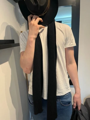 Svart ribbstickad halsduk - En lång, svart halsduk i ribbstickat material som ger en stilren och enkel look. Perfekt att slänga över en t-shirt eller jacka för extra värme och stil. Halsduken har en smal och avlång form och passar till de flesta outfits.