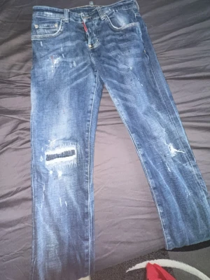 Blå distressed jeans från DSQUARED2 - Säljer ett par blå jeans från DSQUARED2 med slitningar, färgstänk och patchdetalj på ena benet. Klassisk femficksmodell med raka ben och logotyp på bakfickan. Jeansen har en cool, använd look och är tillverkade i denim.