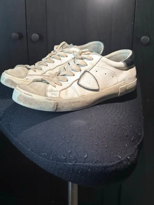 Vita Philippe Model sneakers - Snygga vita sneakers från Philippe Model med grå detaljer och klassisk låg siluett. Skorna har en ikonisk sköld på sidan, vita skosnören och en slitstark gummisula. Materialet är läder med kontrasterande detaljer vid häl och tunga. Perfekta för en avslappnad streetstyle-look.