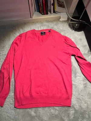 Rosa V-ringad stickad tröja GANT - Snygg rosa stickad tröja från GANT med V-ringning och diskret broderad logga på bröstet. Tröjan har långa ärmar och ribbade muddar vid ärmslut och nederkant. Perfekt för dig som vill ha en klassisk och stilren look.