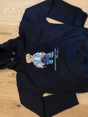 Svart hoodie Polo Bear Ralph Lauren - Svart hoodie från Ralph Lauren med den ikoniska Polo Bear-trycket på bröstet. Hoodien har huva med dragsko och en stor magficka. Materialet ser ut att vara mjukt och bekvämt, perfekt för en avslappnad stil. Texten 'POLO BEAR BY RALPH LAUREN' under björnen.