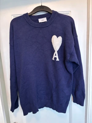 Blå stickad tröja från Ami Paris - Mysig blå stickad tröja från Ami Paris med rund halsringning och ribbade muddar. På bröstet finns ett vitt hjärta och bokstaven A, vilket är signaturdetaljen från märket. Perfekt för dig som gillar stilrena och trendiga plagg.