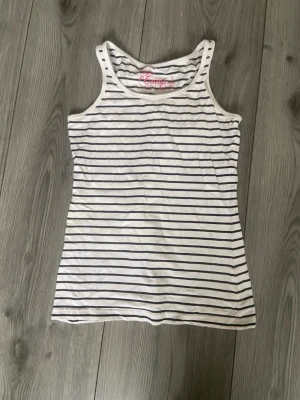 Randig linnetopp från KappAhl - Säljer en vit och marinblå randig linnetopp från KappAhl. Toppen är ärmlös med klassisk rund halsringning och har ett tidlöst, smalt randigt mönster. Perfekt att matcha med jeans eller shorts för en enkel och snygg look.