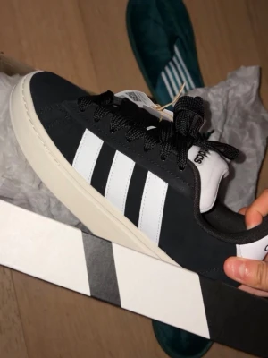 Svarta Adidas sneakers med vita ränder - Klassiska Adidas sneakers i svart mocka med vita läderdetaljer och de ikoniska tre ränderna på sidan. Skorna har svart snörning, vit plös med Adidas-logga och en tjock vit sula. Passar med allt, använda få tal gånger, hör av er om ni har några frågor 🤗