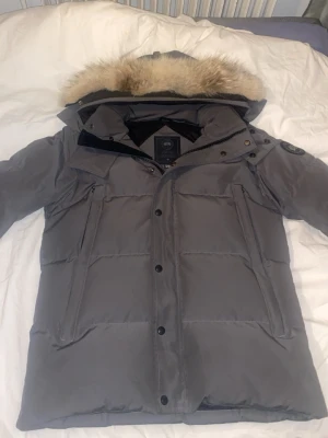 Grå dunjacka från Canada Goose - Snygg grå dunjacka från Canada Goose med avtagbar huva och pälsdetalj. Jackan har klassisk quiltad design, långa ärmar och dragkedja med knappar framtill. Perfekt för kalla vinterdagar och riktigt varm.
