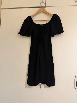 Svart kort puffärmad klänning - Svart klänning med kort längd och puffiga kortärmar. Klänningen har en söt knytning framtill vid bysten och en lätt utställd passform. Perfekt för dig som gillar en enkel men trendig stil.
