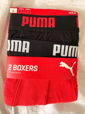 Puma 2-pack boxers röd & svart L - Tvåpack boxerkalsonger från Puma i storlek L. Ena paret är helsvart med röd logotyp i midjan, det andra är helrött med svart resår och vit logotyp. Tillverkade i bomullsstretch för skön passform. Perfekta för dig som gillar sportig stil.