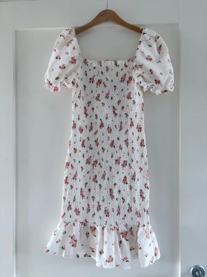 Vit blommig kortklänning med puffärm - Supersöt vit kortklänning med små rosa blommor, puffärmar och volangkant nertill. Klänningen har smockad överdel och är tillverkad i bomull, vilket gör den både bekväm och luftig. Perfekt för sommarens alla tillfällen!