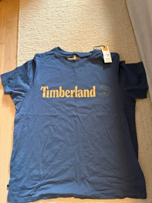 Blå Timberland t-shirt med tryck - Snygg blå t-shirt från Timberland med gul logga och text framtill. Klassisk passform och rund hals. Tillverkad i mjuk bomull som känns skön mot huden. Perfekt för dig som gillar enkel men stilren streetwear.