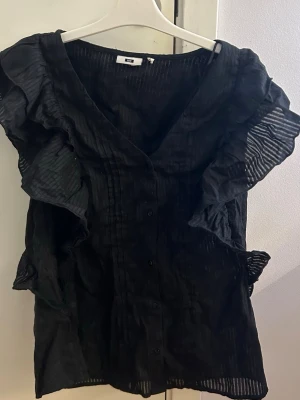 Svart blus med volang - Snygg svart blus från med knappar framtill och stora volanger längs ärmarna. Blusen har v-ringning och är tillverkad i ett tunt, transparent material med subtila ränder. 