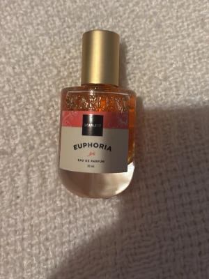 Scarlett Euphoria Eau de Parfum 30 ml - Scarlett Euphoria Eau de Parfum i en rund glasflaska med guldlock. Flaskan rymmer 30 ml och har en etikett i vitt och rosa med blommönster. Doften är elegant och flaskan är smidig att ta med i väskan.