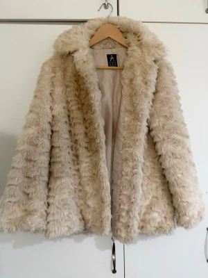 Beige fluffig pälsjacka med krage - Säljer en supermjuk och fluffig beige pälsjacka. Jackan har en bred krage och är helfodrad på insidan. Perfekt för att hålla värmen under kalla vinterdagar och ger en riktigt trendig look.