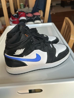 Air Jordan 1 Mid svart/vit/blå - Snygga Air Jordan 1 Mid sneakers i svart och vitt med blå swoosh och orange detaljer på plösen. Klassisk high-top siluett med perforerad tåbox och svart snörning. Tillverkade i läder och textil för en clean och sportig look. Knappt använda , i nyskick 