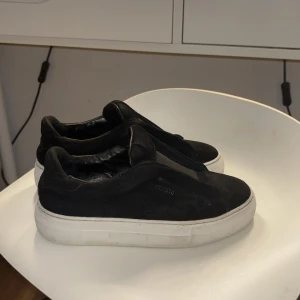 Svarta slip-on sneakers från Arigato - Stilrena svarta slip-on sneakers från Arigato med tjock vit sula. Ovandelen är i mocka och textil, och skon har minimalistisk design utan snörning. Märkeslogga syns på ovansidan. Perfekta för en clean och modern look.