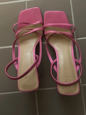 Rosa sandaletter med blockklack H&M - Säljer ett par trendiga rosa sandaletter från H&M med fyrkantig tå och tunna remmar. Skorna har en hög blockklack och stilren design. Tillverkade i syntetmaterial med beige innersula. Perfekta för dig som vill sticka ut med färg.