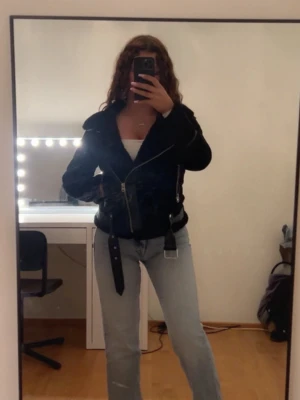 Zara jacka - Snygg svart skinnjacka från Vila med mjukt fuskpälsfoder, bred krage och detaljerade dragkedjor. Jackan har ett bälte i midjan och två snedställda fickor med dragkedja. Perfekt för dig som vill ha en trendig och varm jacka till hösten.