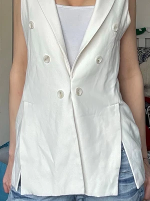 Vit ärmlös kostymväst från Zara - Snygg vit kostymväst från Zara med dubbelknäppning och breda slag. Västen är ärmlös och har två fickor framtill. Materialet är slätt och ger en elegant look. Perfekt att styla över en topp eller skjorta. Skriv gärna om ni har frågor eller vill ha fler bilder!!