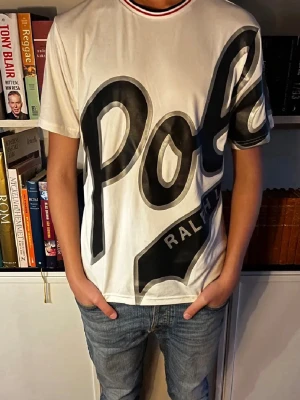 Vit t-shirt med stor Polo-logga - Vit t-shirt från Ralph Lauren med stor svart Polo-logga över hela framsidan och detaljer i grått. T-shirten har korta ärmar och en ribbad krage med röd och svart kant. Perfekt för dig som gillar bolda prints och klassiska märken.