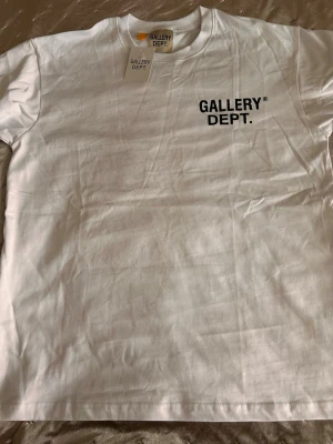 Vit t-shirt från Gallery Dept. - Säljer en clean vit t-shirt från Gallery Dept. med svart logga tryckt på bröstet. Klassisk passform och rund halsringning. Perfekt basic-plagg för dig som gillar streetwear och enkel stil. Storlek M men sitter som L