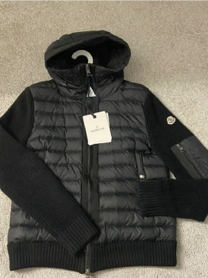 Moncler Cardgian  - Snygg svart pufferjacka från Moncler med stickade ärmar och quiltad framsida. Jackan har huva, dragkedja framtill och Moncler-logga på ärmen. Perfekt för dig som vill ha en stilren och sportig look med exklusiv känsla nu på sena sommarkvällar. Skick stabil 8.5/ 10 Nfc scan funkar. Hör av dig vid minsta lila tvekan så löser vi pris och andes frågor Mvh Emma 💗🤗