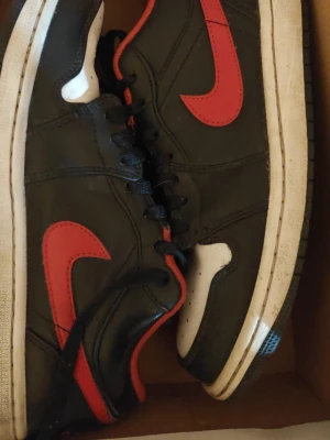 Nike svart-röda sneakers - Säljer ett par klassiska Nike sneakers i svart läder med röda detaljer och vit sula. Skorna har den ikoniska röda swoosh-loggan på sidan och svart snörning. Tåboxen är vit med perforeringar för ventilation. Perfekta för dig som gillar streetwear.