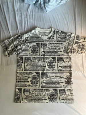 Vit t-shirt med tryck från Levi's - Cool vit t-shirt från Levi's med unikt svart tryck över hela plagget som liknar vintage etiketter. T-shirten har rund hals och korta ärmar, perfekt för dig som vill sticka ut med en retro vibe. 