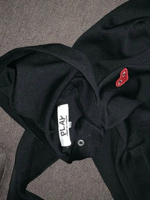 Svart hoodie Comme des Garçons Play - Svart hoodie från Comme des Garçons Play med det ikoniska röda hjärtat med ögon broderat på bröstet. Klassisk passform med huva och ribbade muddar. Perfekt för dig som gillar streetwear och vill ha en stilren men ändå unik look.
