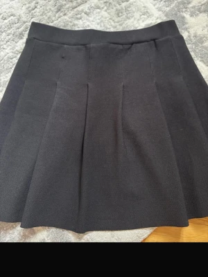 Svart plisserad kortkjol - Snygg svart kortkjol med plisserade veck och bred linning. Kjolen har en klassisk och stilren look som passar till många olika outfits. Materialet känns mjukt och följsamt, perfekt för dig som gillar enkel men trendig stil.