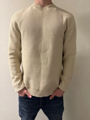 Beige stickad tröja med hög krage - Säljer en stilren beige stickad tröja med högre ribbad krage och långa ärmar. Tröjan har en enkel, rak passform och är tillverkad i ett mjukt, grovt stickat material. Perfekt för lager på lager och passar till jeans eller chinos. Ny pris 600 men säljs endast för 400kr 