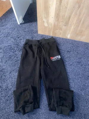 Svarta Trapstar mjukisbyxor - Säljer ett par svarta mjukisbyxor från Trapstar med trycket 'Trapstar It's a Secret' i vitt och rött på vänster ben. Byxorna har resår i midjan med dragsko och muddar vid bensluten. Perfekta för en chill och streetwear-inspirerad look. Hör av er vid frågor eller funderingar 