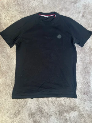 Svart Moncler t-shirt  - Snygg svart t-shirt från Moncler med klassisk rund hals och korta ärmar. T-shirten har en diskret Moncler-logga på bröstet och är tillverkad i mjuk bomull för en bekväm känsla. Perfekt till jeans eller shorts för en clean look. Tröjan är L i storlek men den sitter som en M