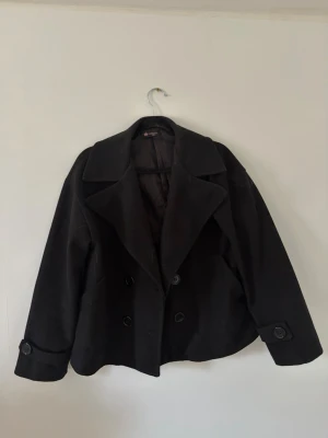Kort svart dubbelknäppt kappa - Stilren svart rock med breda slag och dubbelknäppning framtill. Kortare modell med dekorativa knappar vid ärmsluten och klassisk krage. Perfekt för dig som gillar en tidlös look ny till våren. Obs! Är ett litet hål i fodret vilket ni ser i bild 3. Detta går och fixa enkelt med nål och tråd🥰
