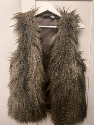 Brun långhårig fuskpälsväst - Säljer en brun långhårig fuskpälsväst med öppen front och mjukt innerfoder. Västen har en naturlig, lite vild look och är perfekt för att addera en trendig touch till din outfit. Märke och storlek saknas på etiketten.