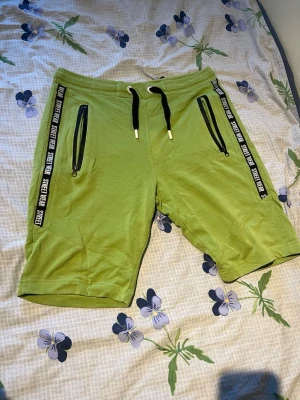 Gröna shorts med snörning och dragkedja - Snygga gröna shorts med svarta snören i midjan och dragkedjeförsedda fickor. Svarta band med texten 'STREET WEAR' längs sidorna ger en sportig vibe. Perfekta för sommaren och avslappnade dagar.