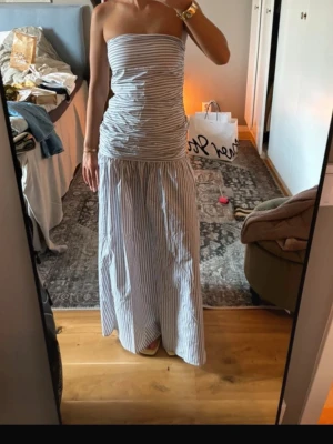 Randig axelbandslös långklänning - Säljer en snygg axelbandslös långklänning med smala vertikala ränder i vitt och grått. Klänningen har en figurnära överdel och ett längre, löst fallande kjolparti. Perfekt för sommarens alla tillfällen!