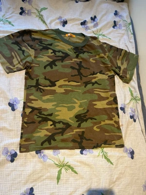Två camo t-shirts i bomull - Säljer två snygga t-shirts med camouflage-mönster. Ena t-shirten har klassiskt grönt camo och den andra har mörkare toner i svart och grått. Båda är kortärmade och gjorda i mjuk bomull, perfekta för en avslappnad streetwear-look.