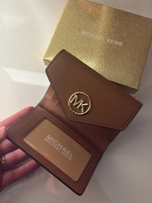 Michael Kors brun och beige plånbok - Snygg plånbok från Michael Kors i brunt och beige med MK-logga i guld framtill. Plånboken har kuvertformad flik, fack för kort och ID-fönster. Tillverkad i läder och canvas med klassiskt MK-mönster. Perfekt för dig som vill ha stilren och trendig accessoar.