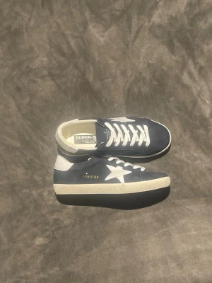 Blåa sneakers från Golden Goose - Snygga blå sneakers från Golden Goose med vit stjärndetalj på sidan och vita snören. Skorna har en klassisk låg profil och är tillverkade i skinn med kontrasterande vit häl och sula. Perfekta för en trendig och avslappnad stil.