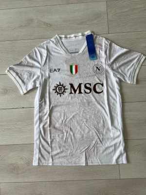 EA7 Napoli Jersey,vit - Vit Napoli-tröja från EA7 – riktigt clean piece som sticker ut på ett subtilt sätt. Snygga detaljer i tyget och fet front med MSC-loggan. Helt ny med tags kvar. Storlek S. Perfekt om du vill ha något som känns både exklusivt och enkelt 🔥