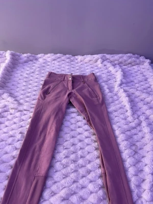 Lila ridbyxor med guld-detaljer - Säljer ett par lila ridbyxor från Pikeur med raka ben och snygga guld-detaljer på fickorna. Byxorna har två framfickor med dragkedja och två bakfickor, varav en har en broderad logga i guld. Materialet är stretchigt och bekvämt.