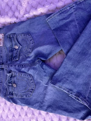 Blå True Religion Joey flare jeans - Säljer ett par blå Joey low rise flare jeans från True Religion. Jeansen har snygga kontrastsömmar, stora fickor med lock och silverfärgade knappar. Materialet är klassisk denim och passformen är utsvängd nertill för en retro vibe.