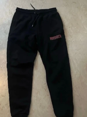Svarta mjukisbyxor från Hoodrich - Svarta mjukisbyxor från Hoodrich med röd logga på vänster lår. Byxorna har dragsko i midjan och muddar vid bensluten. Perfekta för en avslappnad och street-inspirerad stil.