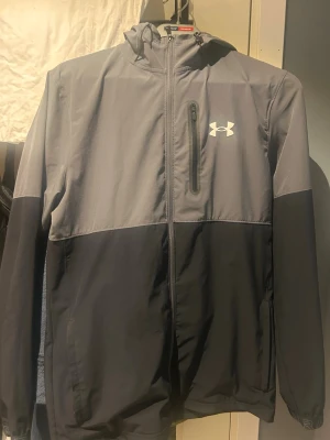 Grå och svart vindjacka Under Armour - Snygg tvåfärgad vindjacka från Under Armour i grått och svart. Jackan har huva, hel dragkedja framtill, bröstficka med dragkedja och logga på bröstet. Tillverkad i lätt och vindtätt material, perfekt för blåsiga dagar. Finns ej att köp någonstans längre!!!