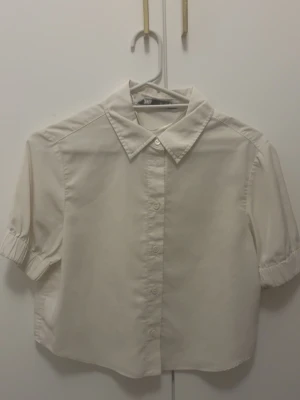 Vit kortärmad blus från Zara - Stilren vit blus från Zara med klassisk krage och knäppning framtill. Blusen har korta ärmar med lätt puff och resår vid ärmslut. Passar perfekt till både jeans och kjol för en fräsch look.