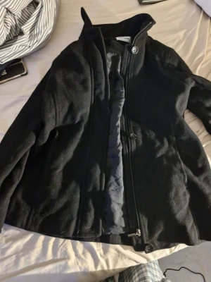 Svart ullrock med hög krage - Stilren svart rock i ullblandning med hög krage och dragkedja samt knappar framtill. Rocken har en rak passform och diskreta sömmar för en clean look. Perfekt för kyliga dagar när du vill hålla stilen enkel och snygg.