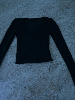 Svart omlottopp med lång ärm - Snygg svart omlottopp med lång ärm och v-ringad hals. Toppen har en croppad passform och är tillverkad i ett mjukt, stretchigt material som känns skönt mot huden. Perfekt att styla med höga jeans eller kjol.