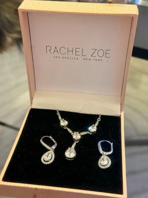Rachel Zoe smyckesset med kristaller - Snyggt smyckesset från Rachel Zoe med matchande halsband och örhängen i silverfärg. Både halsbandet och örhängena har droppformade hängen med gnistrande klara kristaller. Perfekt för dig som gillar elegant och stilren design.