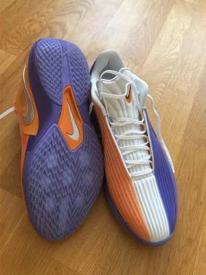 Nike basketskor lila och orange - Säljer ett par riktigt snygga Nike basketkor i lila, orange och vitt. Skorna har en nice design med ribbad ovandel, vit snörning och tydlig Swoosh-logga på sidan. Sulan är lila med orange detaljer och greppvänligt mönster. Perfekta för dig som gillar färg.