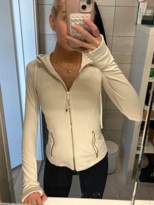 Lululemon Hooded Define Vit och Guld - Snygg och figurnära träningsjacka från Lululemon i ljus ivory/vit färg. Jackan har huva, gullet dragkedja framtill, två fickor med dragkedja och tumhål vid ärmsluten. Tillverkad i stretchigt funktionsmaterial som andas. Strl 2. Köpt i London för 1500. Använd 1-2 gånger.