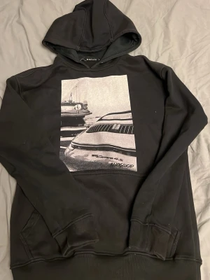 Svart hoodie från Blinglucid - Svart hoodie från Blinglucid med stor grafisk print framtill som visar bilar i svartvitt. Klassisk huva och känguruficka. Tillverkad i mjukt material som känns skönt mot huden. Perfekt för dig som gillar streetwear och unika prints.